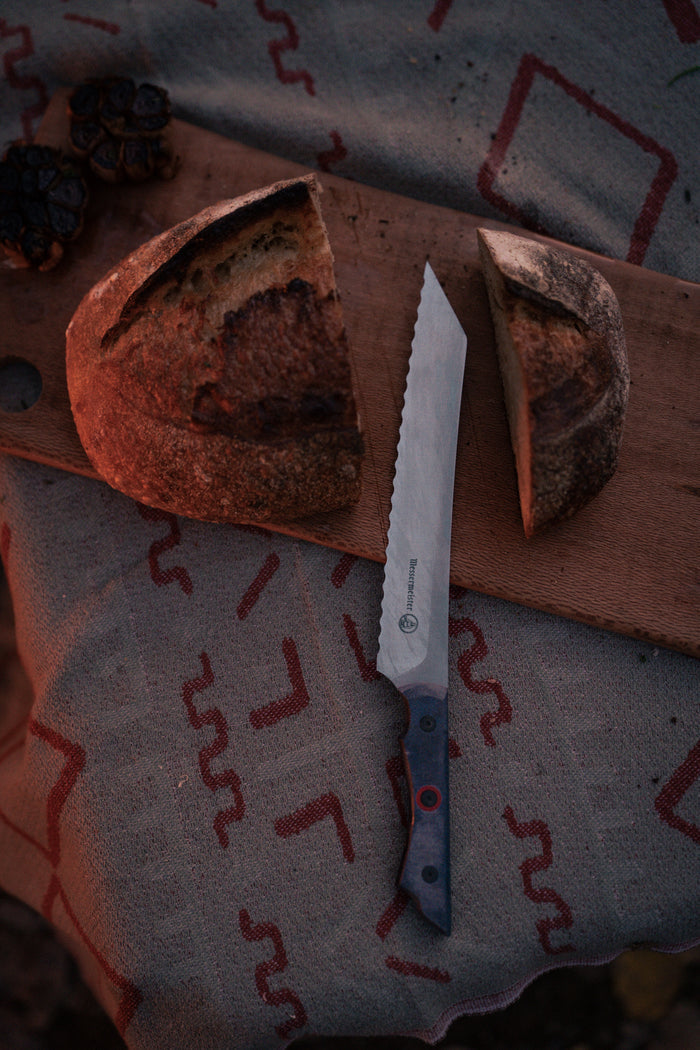 A serrated knife on a patterned cloth beside a sliced loaf of bread and roasted garlic on a wooden board.