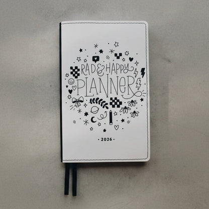 A white 2026 'Rad & Happy Planner' with black doodle illustrations and ribbon bookmarks, resting on a textured gray surface.