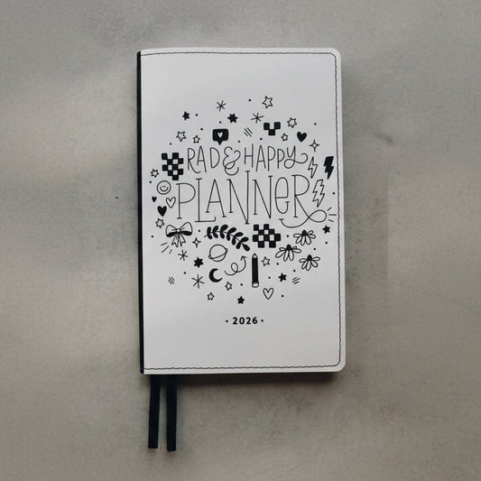 A white 2026 'Rad & Happy Planner' with black doodle illustrations and ribbon bookmarks, resting on a textured gray surface.