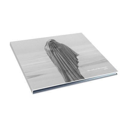A minimalist album cover for The End Of The Ocean, showing a black and white figure draped in fabric on a beach. "the end of the ocean" and "aire" appear in lowercase in the bottom right, evoking quiet redemption.