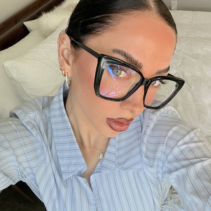 lifestyle - emily gordon brunette influencer wearing light blue cotton blouse with oversized black cat eye blue light glasses