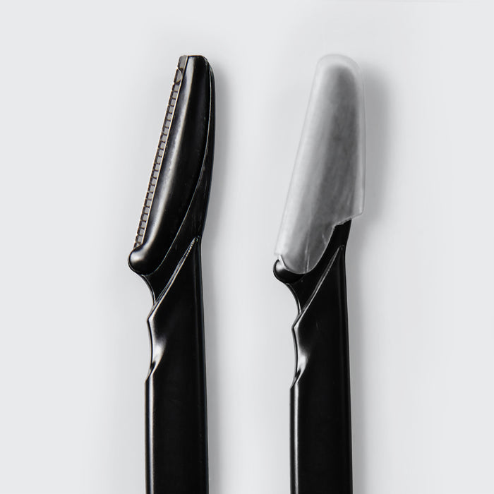 Close-up of two Kitsch Black Dermaplaner blades, one with its protective cover on and one uncovered, on a white background