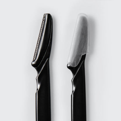 Close-up of two Kitsch Black Dermaplaner blades, one with its protective cover on and one uncovered, on a white background
