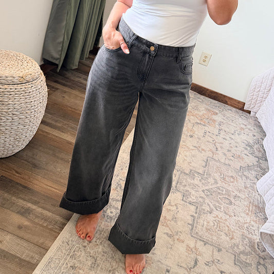 A person wearing dark gray wide-leg cuffed jeans and a white top, standing barefoot on a patterned area rug in a bedroom setting.