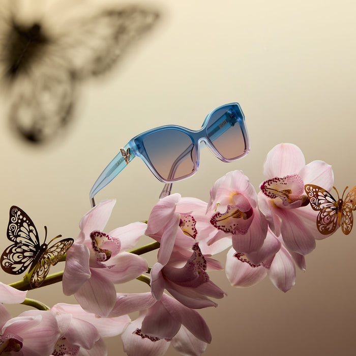 lifestyle - wicked x diff eyewear featuring the glinda upland butterfly cat eye sunglasses with a blue pink gradient frame and blue to pink lenses floating over pink flowers