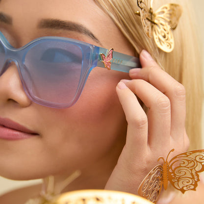 lifestyle - female wearing wicked x diff eyewear featuring the glinda upland butterfly cat eye sunglasses with a blue pink gradient frame and blue to pink lenses detailed view of temple with butterfly decoration