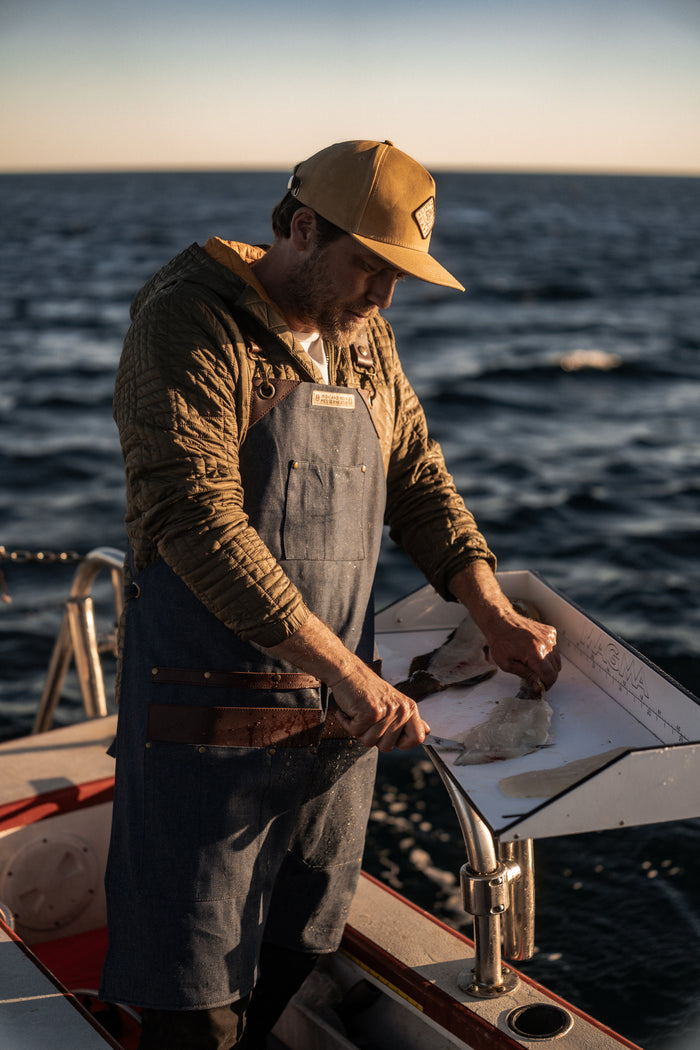 Man in a cap and apron fillets fish on a boat at sunset, with the ocean in the background.