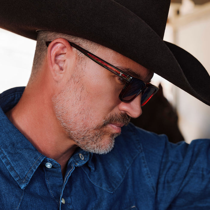 lifestyle - male in horse stables wearing yellowstone x diff eyewear john dutton rectangular sunglasses with a redwood frame and grey polarized lenses side view