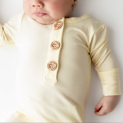 Baby wearing a light yellow-colored outfit with buttons and a matching hat on a white background