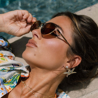 lifestyle - jessie james decker female laying near a swimming pool wearing diff eyewear limoncello oval sunglasses with a gold frame and brown lenses side view