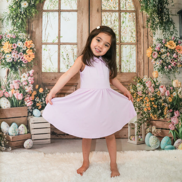 Smiling girl standing in front of a wooden window with flowers holding the hem of the skirt of the Polo Bodysuit Dress in Thistle
