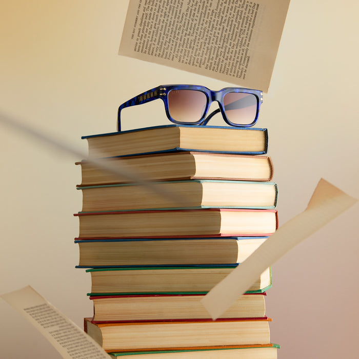 lifestyle - wicked x diff eyewear featuring the prince fiyero square sunglasses with a regal blue tortoise frame and brown gradient lenses laying on a stack of books