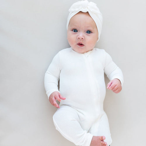 Baby laying on off-white surface wearing Kyte Baby Ribbed Zippered Romper in Cloud and matching Kyte Baby Headwrap
