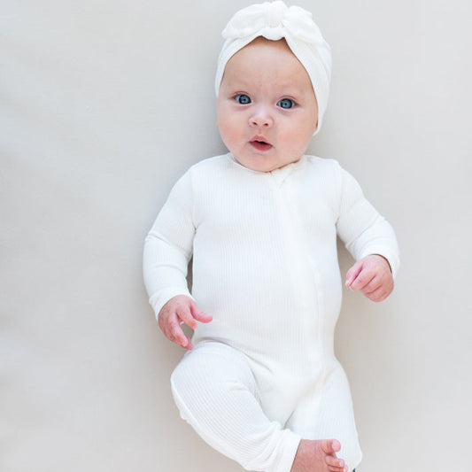 Baby laying on off-white surface wearing Kyte Baby Ribbed Zippered Romper in Cloud and matching Kyte Baby Headwrap