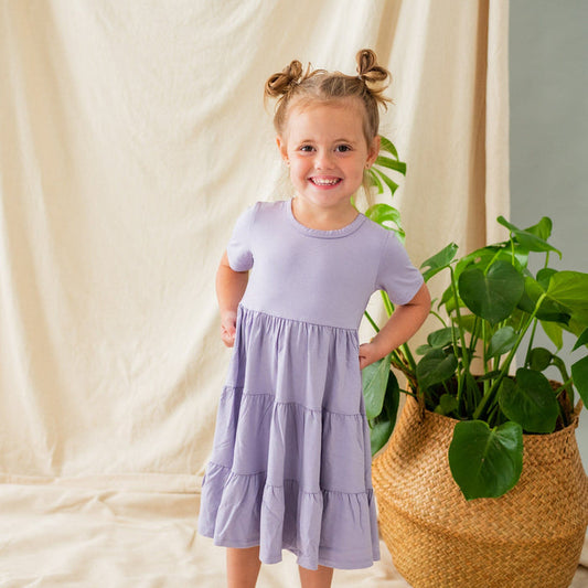 Smiling child standing next to a plant wearing a Short Sleeve Tiered Dress in Taro