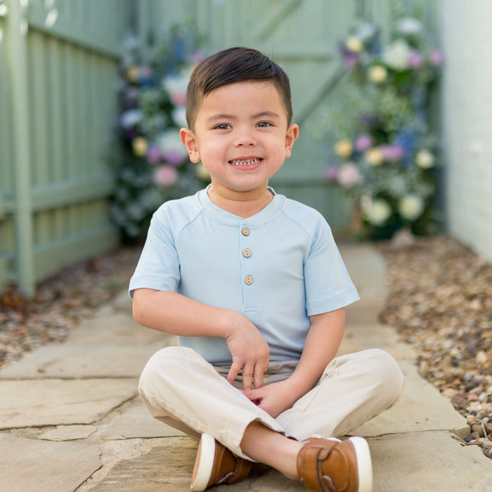 Young boy sitting on a stone path outside wearing the Short Sleeve Toddler Henley Tee in Breeze with khaki colored pants