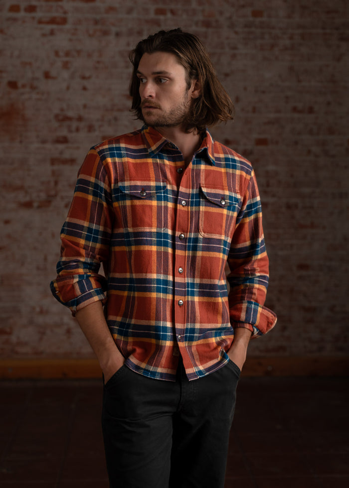 Man in a plaid shirt stands against a brick wall, hands in pockets, gazing to the side with a thoughtful expression.