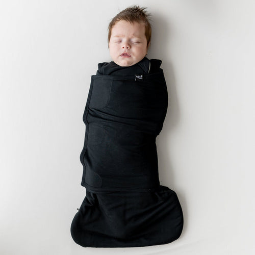 Sleeping infant laying on a light neutral blanket swaddled in the Sleep Bag Swaddler in Midnight 1.0
