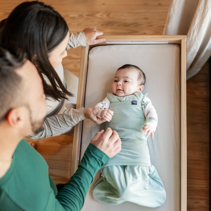 Baby in bassinet while wearing Sleep Bag Swaddler in Sage. Parents are standing next to bassinet, looking down at baby and mother is hold its hand