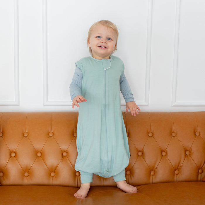 Toddler wearing Kyte Baby Sleep Bag Walker in Sage 0.5 while standing on a couch