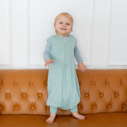 Toddler wearing Kyte Baby Sleep Bag Walker in Sage 0.5 while standing on a couch
