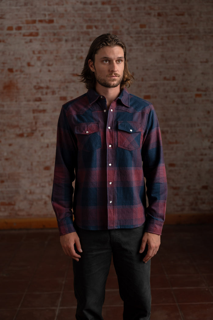 Man in a plaid shirt stands against a rustic brick wall, looking thoughtful. His casual attire contrasts with the textured backdrop.