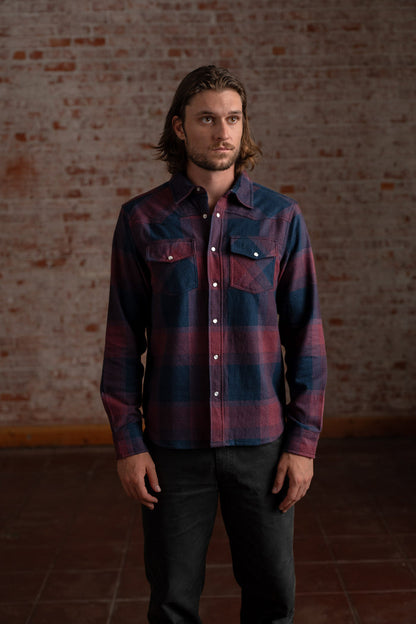 Man in a plaid shirt stands against a rustic brick wall, looking thoughtful. His casual attire contrasts with the textured backdrop.