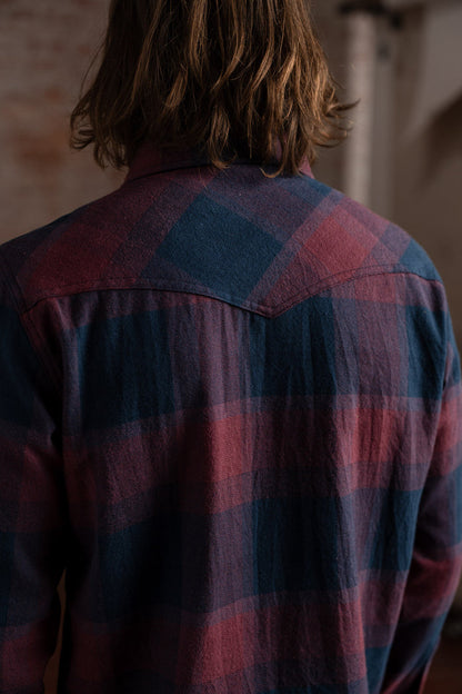 Back view of a person with long hair wearing a red and blue plaid shirt, standing indoors against a blurred background.