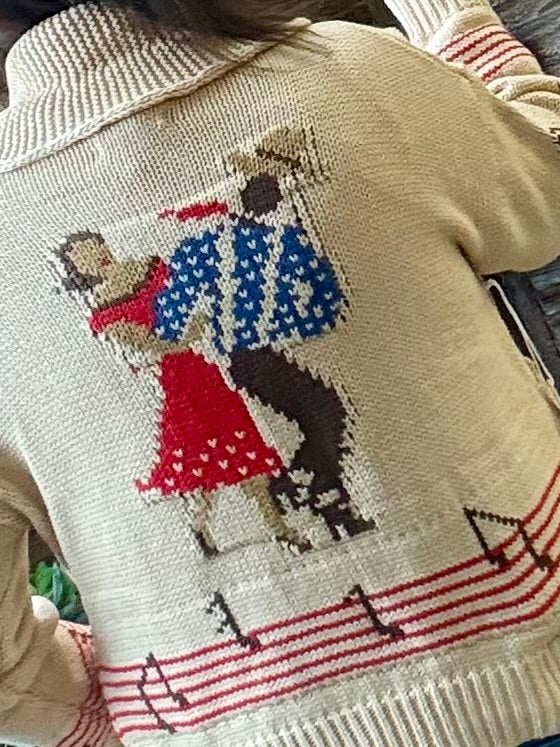 Cream knit cardigan back with blue and red square-dance figures and red striped hem