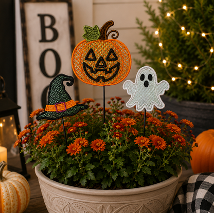 Halloween-themed floral garden stake decorations featuring a jack-o'-lantern, a witch hat, and a ghost, displayed in a pot of orange mums with decorative pumpkins and a lit Christmas tree in the background.