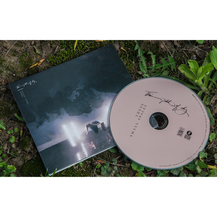 A CD and its case rest on grass. The case shows a dark scene with a car and lights. The CD, labeled Three Small Spaces, has handwritten text and signatures, suggesting it’s by This Patch of Sky, possibly a Hiraeth or Pale Lights collab.