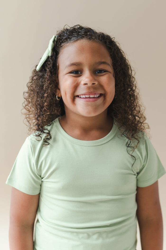 Young girl smiling wearing the Toddler Basic Tee in Basil with a matching alligator clip bow