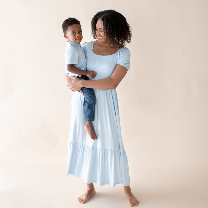Woman in a light blue dress holding a child  in a matching blue polo against a beige background