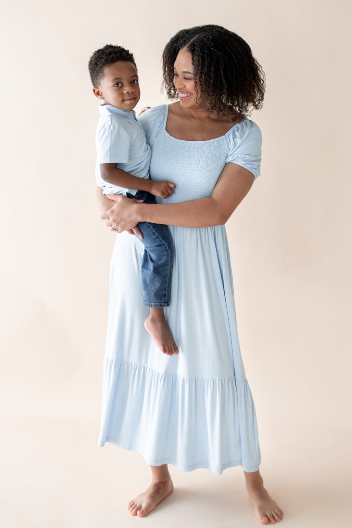 Woman in a light blue dress holding a child  wearing matching blue polo against a beige background
