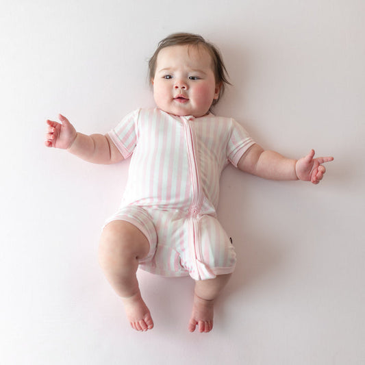 Infant laying on a light pink surface wearing the buttery soft Zipper Shortall Romper in Small Sakura Stripe